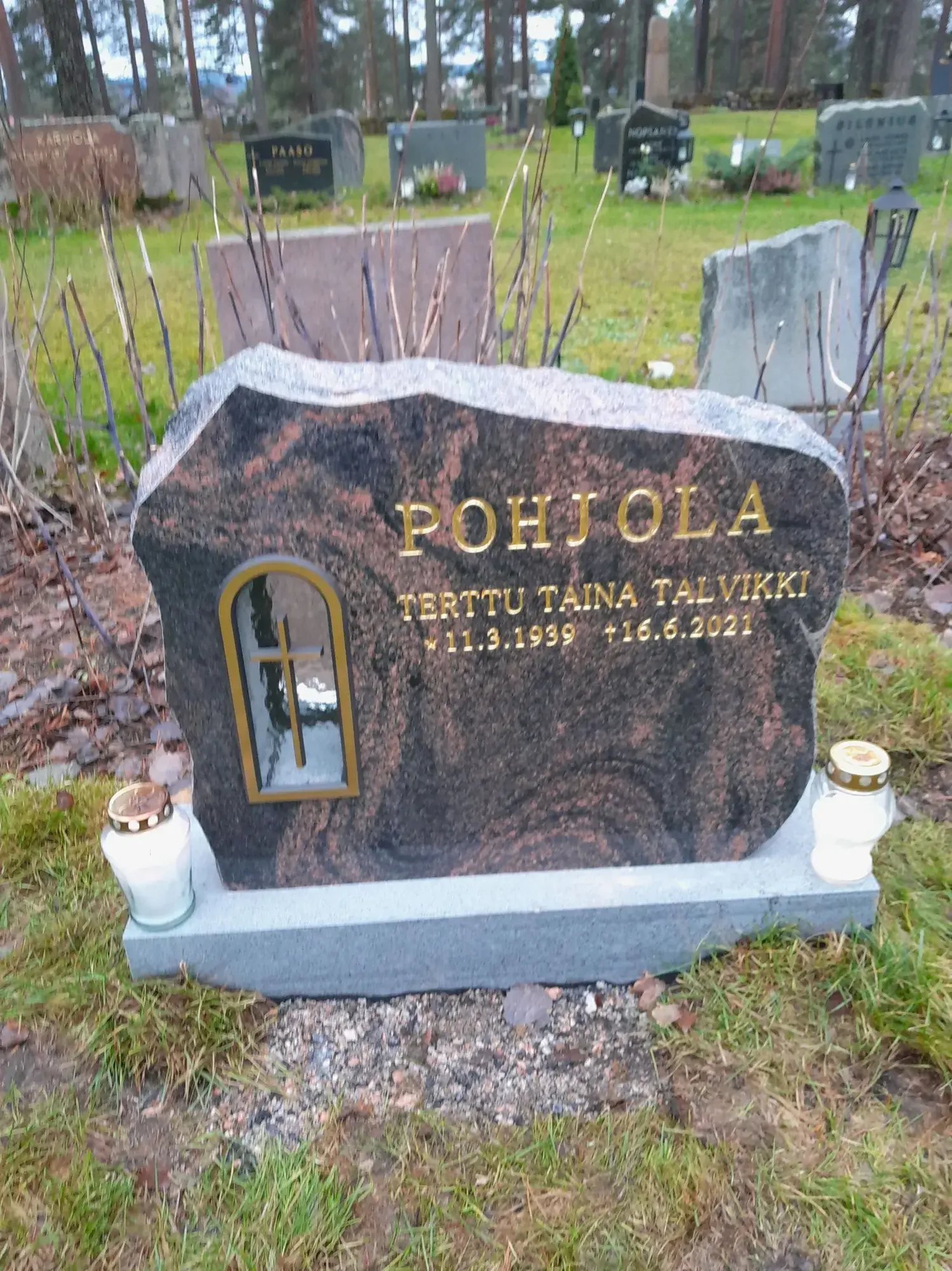 Tummanruskea graniittinen hautakivi, jossa on kultaiset kirjaimet 'POHJOLA', 'TERTTU TAINA TALVIKKI' ja päivämäärät '*11.3.1939 +16.6.2021'. Kiven vasemmassa alaosassa on kultareunainen ikkunanmuotoinen kaiverrus, jossa on risti. Kiven edessä on kaksi valkoista hautakynttilää. Taustalla näkyy vihreää ruohoa, paljaita pensaita, muita hautakiviä ja metsäisiä puita pilvisen taivaan alla.