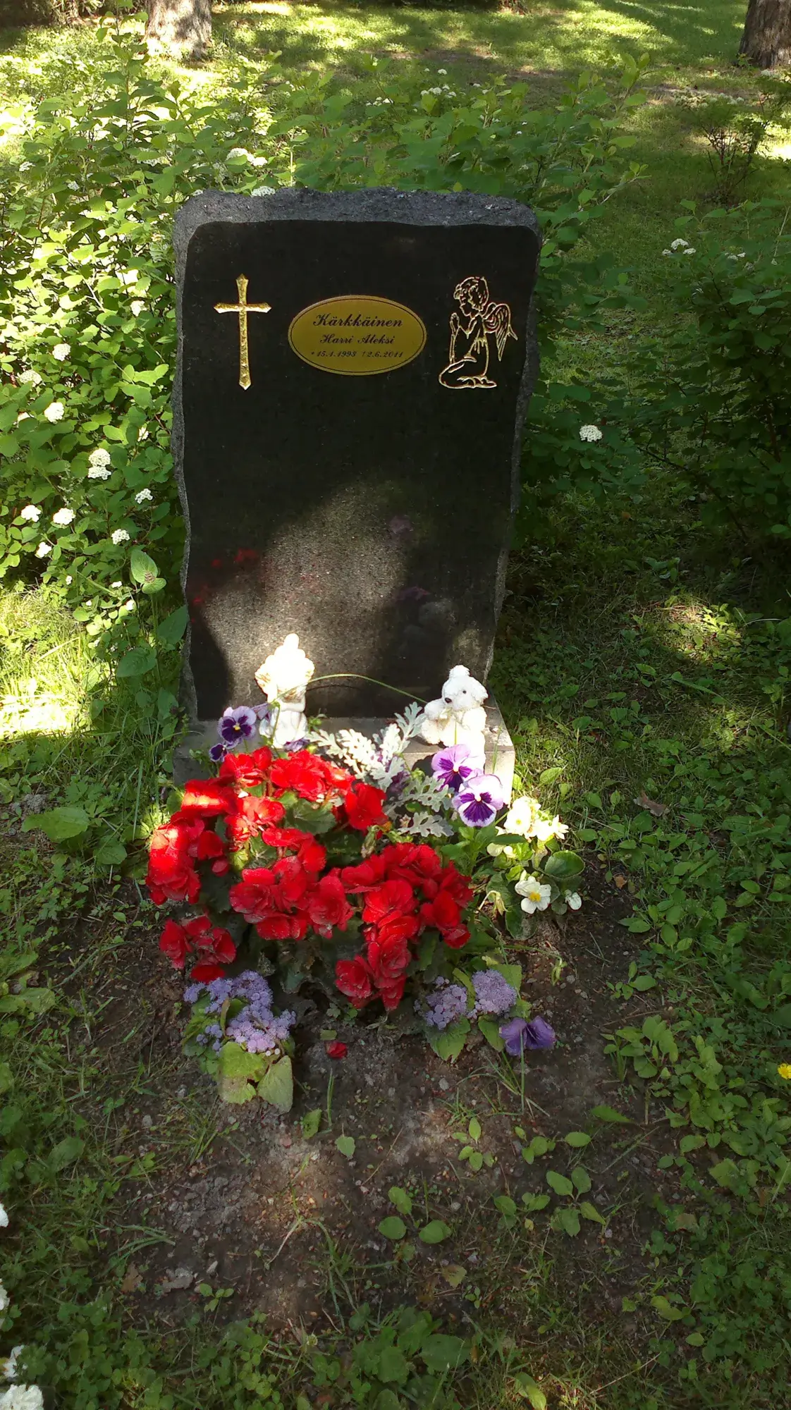 Musta hautakivi, jossa on kultainen risti, soikea nimilaatta ja rukoileva enkeli. Laatan teksti on Kärkkäinen, Harri Aleksi, 15.1.1993 12.6.2011. Kiven edessä on punaisia begonioita, violetteja ja valkoisia orvokkeja, pieni valkoinen patsas ja nalle. Ympärillä on vihreää ruohoa ja pensaita aurinkoisessa valossa.