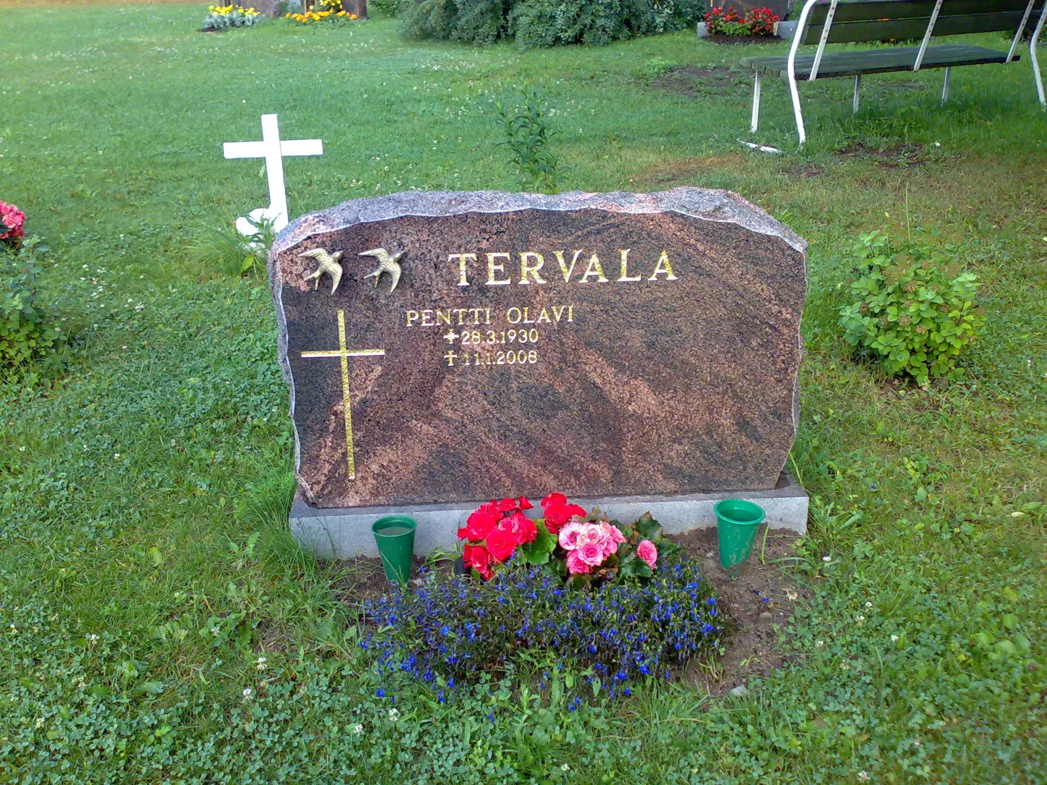Hautakivi vihreällä nurmikolla. Tummanruskeassa graniittikivessä on kultaisin kirjaimin nimi 'TERVALA' ja 'PENTTI OLAVI' sekä syntymä- ja kuolinpäivämäärät '+ 28.3.1930 † 1.1.2008'. Nimen yläpuolella on kaksi kultaista pääskystä ja päivämäärien vieressä kultainen risti. Hautakiven edessä on punaisia, vaaleanpunaisia ja sinisiä kukkia, ja sen molemmin puolin on vihreät kukka-astiat. Taustalla näkyy valkoinen risti ja puinen penkki.