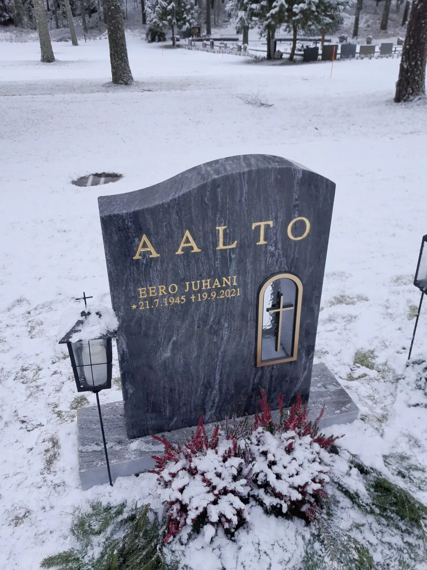 Lumipeitteisellä hautausmaalla etualalla tummanharmaa graniittinen hautakivi. Kivessä on kultaisin kirjaimin nimi 'AALTO', 'EERO JUHANI' sekä syntymä- ja kuolinajat. Oikealla puolella on syvennys, jossa on kultareunainen kaari ja kultainen risti. Hautakiven vasemmalla puolella on lumen peittämä musta lyhty, jonka päällä on risti. Hautakiven edessä on lumen peittämiä punaisia kanervia ja vihreitä männynoksia. Taustalla näkyy lumisia puita ja muita hautakiviä pilvisessä säässä.