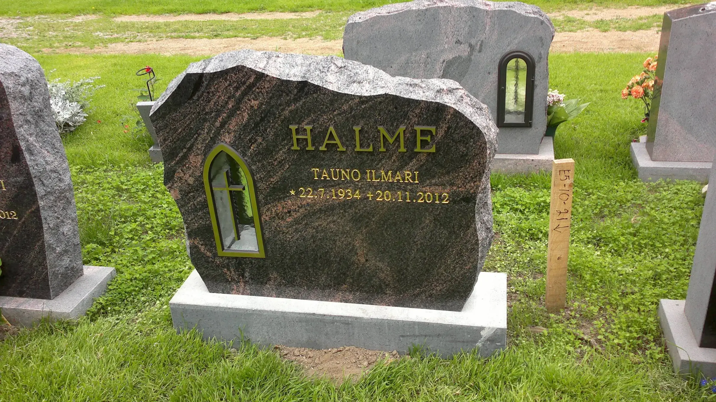 Tumma graniittinen hautakivi, jossa on kultaiset kaiverrukset 'HALME TAUNO ILMARI * 22.7.1934 + 20.11.2012'. Hautakivessä on myös kultareunainen kaareva syvennys, jonka sisällä on risti. Kivi seisoo vaaleanharmaalla alustalla vihreällä nurmikolla. Oikealla on puinen paalu merkinnällä 'E5-10-21V' ja taustalla muita hautakiviä sekä oransseja kukkia kirkkaassa päivänvalossa.