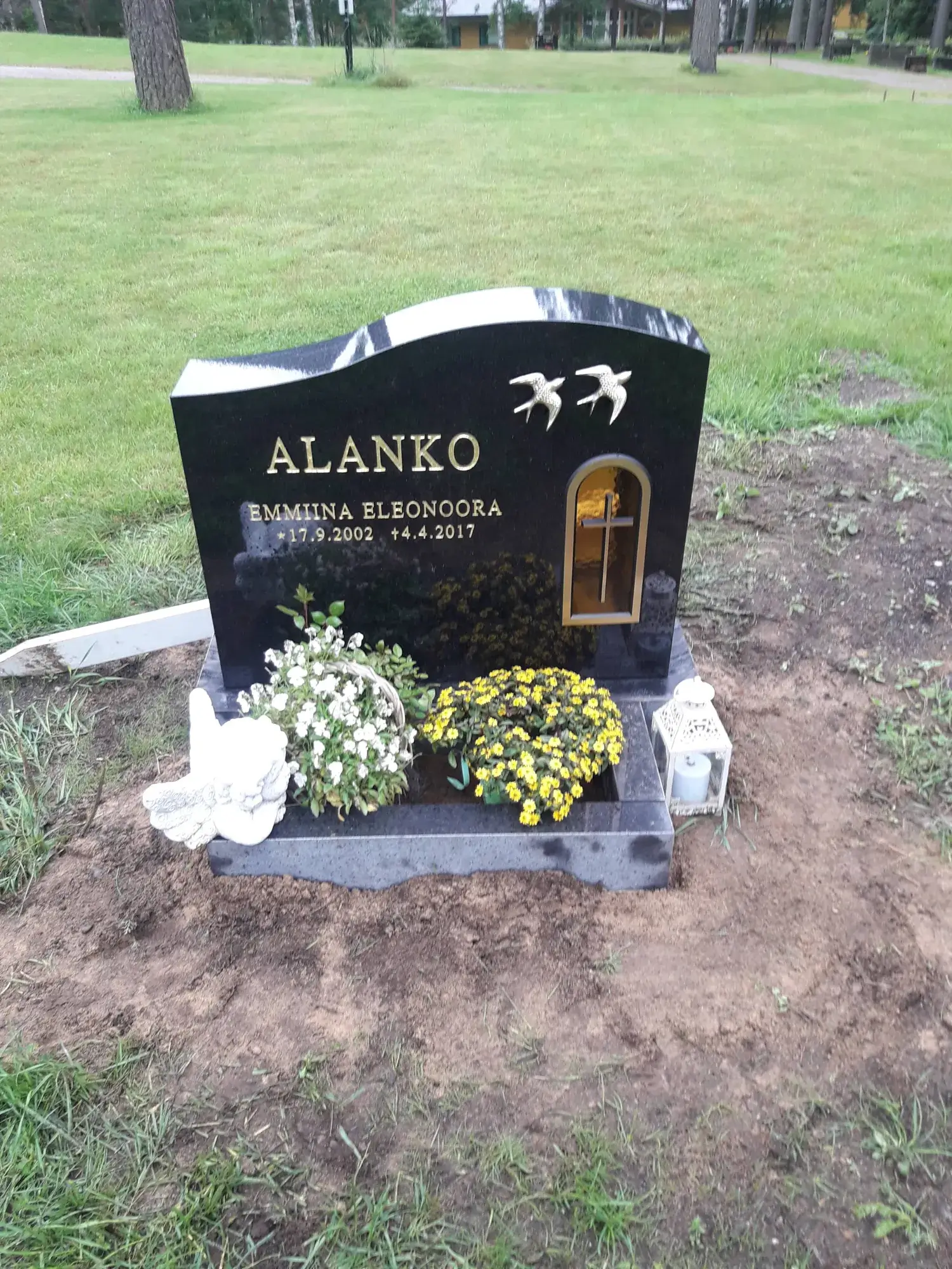Musta graniittinen hautakivi, jossa on kultaisin kirjaimin nimet 'ALANKO' ja 'EMMIINA ELEONOORA' sekä syntymä- ja kuolinpäivät 17.9.2002 ja 4.4.2017. Kiven yläosassa on kaksi kultaista pääskyä ja oikealla puolella kultainen kaari-ikkuna, jossa on risti. Hautakiven edustalla on valkoinen enkelihahmo, korissa valkoisia kukkia, keltainen kukkaseppele ja valkoinen lyhty kynttilällä. Taustalla näkyy vihreää nurmikkoa ja puita hautausmaa-alueella pilvisenä päivänä.