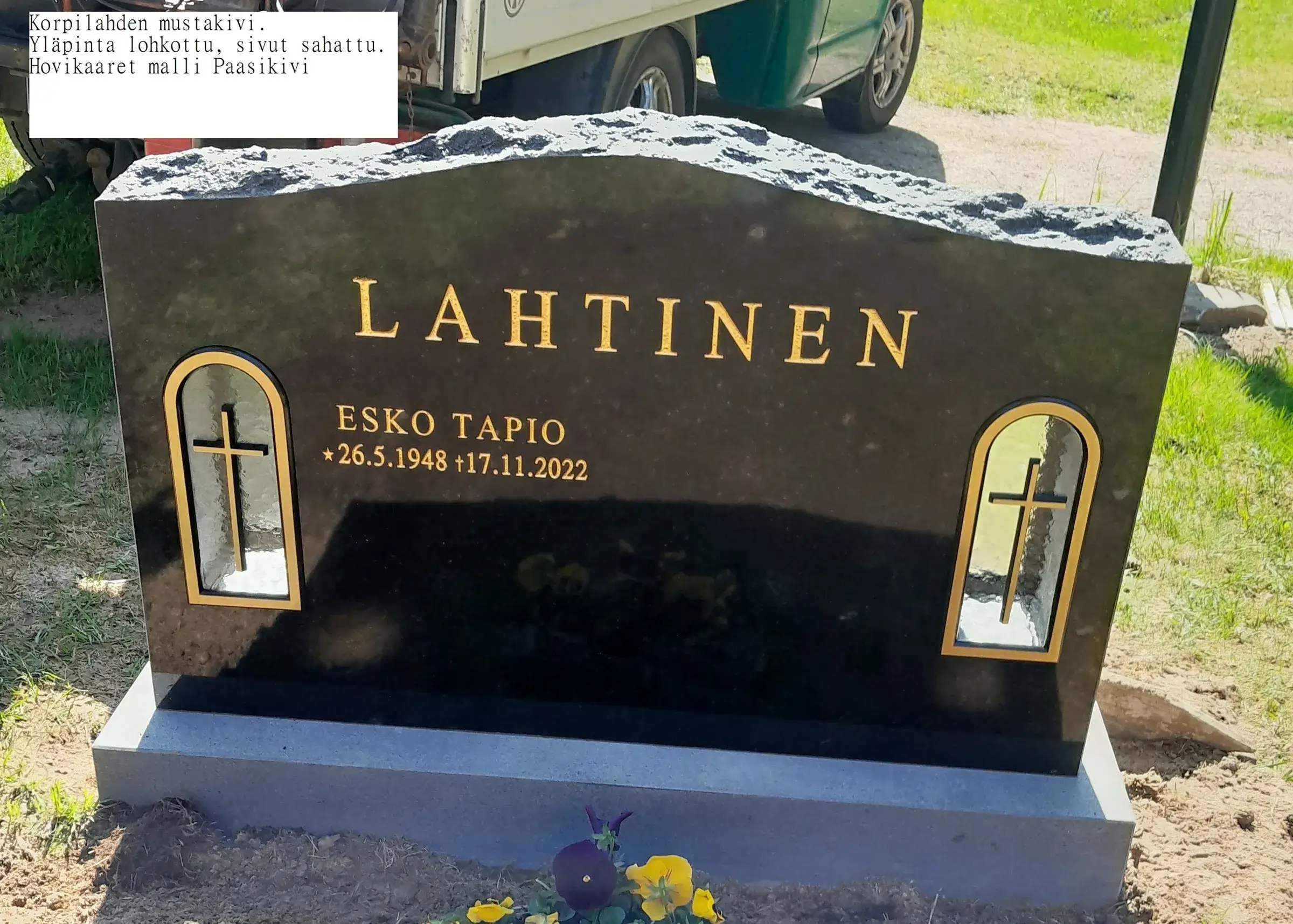 Musta graniittinen hautakivi, jossa kultaiset kirjaimet "LAHTINEN", "ESKO TAPIO" ja päivämäärät "★ 26.5.1948 † 17.11.2022". Kiven yläpinta on luonnonlohkottu ja sivut sahattu. Molemmilla puolilla on kaarevat syvennykset, joissa on kultainen risti. Hautakivi seisoo vaaleanharmaalla jalustalla vihreän ruohon ja mullan keskellä. Alhaalla on muutamia keltaisia ja violetteja kukkia. Taustalla näkyy osittain ajoneuvo ja hiekkatie aurinkoisessa valossa.